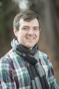 Photo of man, smiling, clean shaven. He has a short brown hair turned to one side with flicks of gray. It's taken in the fall, as he wears a black and gray scarf and the autumn leaves come through in the background.